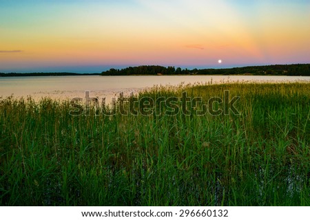 Full moon over lake in late evening. Reeds in foreground and distant cabins on opposite shore. Calm water and fine sky.