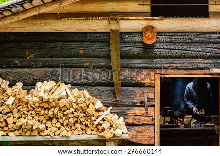 Woodsman cooking over open fire inside a cabin. Firewood and hand saw outside cabin. Walls are burnt instead of painted.