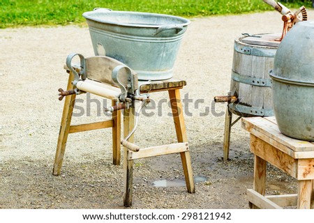 Old fashioned mangle with washing tub on top and crank at side. Several old laundry utensils partly visible to the right. Ground is wet gravel with small puddle under mangle. No person visible.