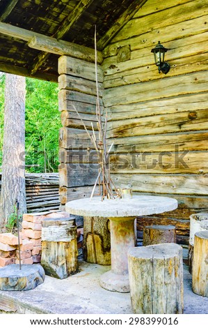 Outdoor furniture made of cut timber, concrete and other materials. Furniture is placed on a porch outside a cabin in the woods. Cabin is timbered with unpainted logs. Forest visible to the left.