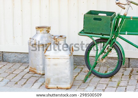 Green vintage bicycle with luggage crate in front to hold merchandise and goods. Two metal milk bottles on the ground in front of it. This has a real retro feel to it. Street is made of granite stone.