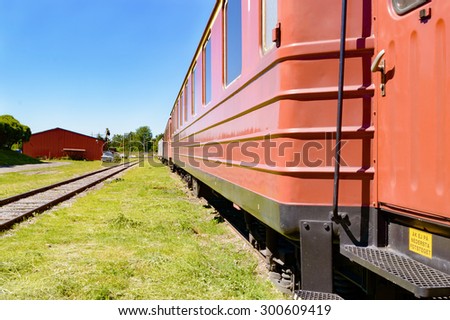 Line of passenger train cars standing on railroad track. Blue sky above and green grass cover the ground beside the cars. Cars are mostly red. No person visible.