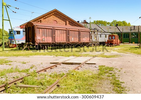 Abandoned trains and cars outside an old service depot with railroad tracks leading up to building. Small railway crossing in foreground. No person visible.