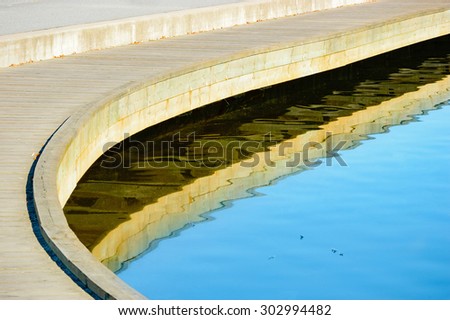 Bending walkway hovering above water. Some feathers float in the blue, calm water. Fine reflections from underneath the walkway. Copyspace in water.