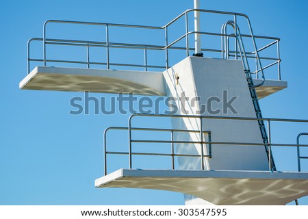 A white diving board or tower against a clear blue sky. Reflections are seen underneath. No person visible.