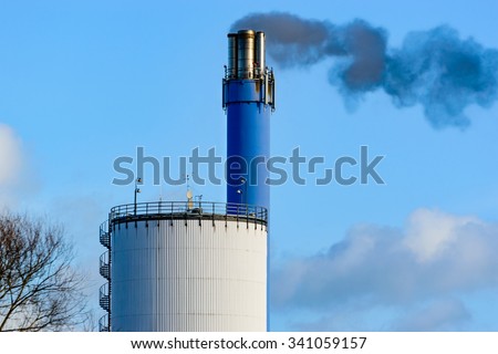 Dark, black smoke is coming out of a blue chimney. Some kind of silo or storage building is seen in front. Blue sky in background. Some clouds drift by.