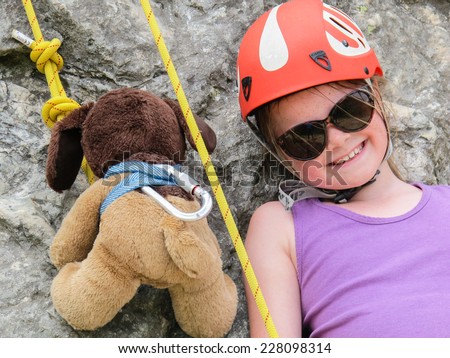Girl in a climbing helmet about to rock climb with her teddy soft toy attached to a climbing rope next to her.