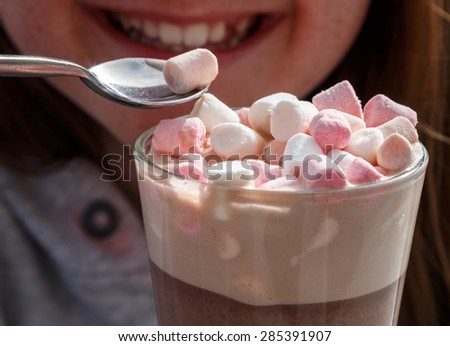 Marshmallows on the top of a hot chocolate drink in a glass being eaten by a young smiling pretty girl.  She is smiling as she picks up a marshmallow with a spoon.