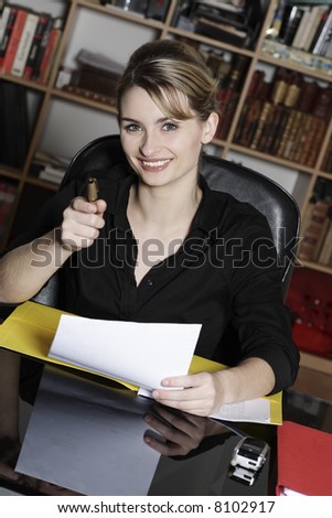 cute and smiling businesswoman at the office desk pointing his pen as his finger at you