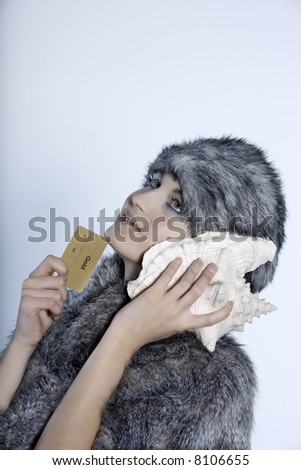 studio shot portrait of a beautiful woman russian type in a fur coat and hat holding a credit card  and listening to the sea through a shell
