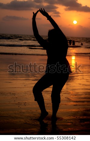 A silhouette of a woman in a traditional Indian Classical dance pose, on a beach at sunset.