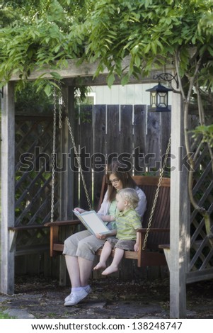 A mother and her two-year old son enjoy reading a book together while sitting on a swing in an arbor. Room around image for text