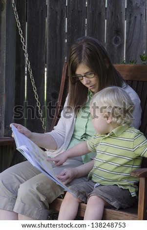 A toddler points to something in a book as he sits with  his mother on a wooden swing