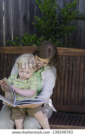 A toddler turns the page of a book as he sits on his mother's lap on a wooden bench swing.