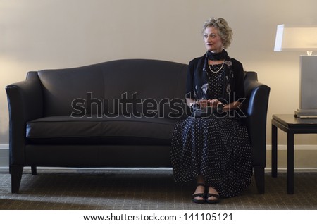 A solitary senior woman sits shoved against one arm rest as though for protection. She looks apprehensive, perhaps in anticipation of someone she\'s there to meet. Possibly a recent widow.