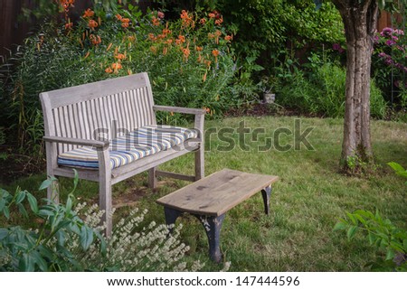 A weathered wooden bench with faded cushion and a rustic table sit invitingly in a secluded garden nook with a background of orange tiger lilies.