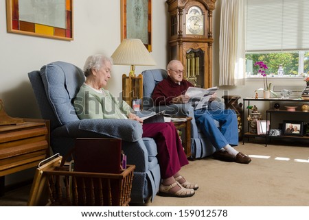 An elderly couple quietly enjoy each others company as they read together in their comfortable living room.