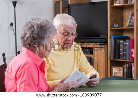 An elderly couple in their comfortable home work together as they read instructions for their new phone.