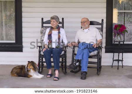 An elderly married couple in their eighties watch the world go by from their porch with their sheltie dog by their side.