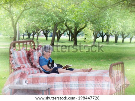 A senior woman enjoys quiet pleasure reading with her dog as she relaxes on an old-fashioned bed placed at the edge of a walnut grove as a private summer retreat.