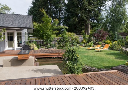 Back yard of a modern Pacific Northwest home featuring a deck spanning a creek-like water feature with a landscaped lawn and two inviting lawn chairs in the background.