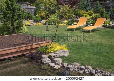 A pair of bright yellow designer lawn chairs invite you to relax in this beautifully landscaped backyard with deck spanning a creek in the foreground.