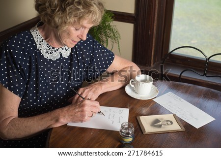 In a historical scene from the 1930Ã?Â¢??s or 1940Ã?Â¢??s, an attractive elderly woman writes a letter as she sits at a table by a window.