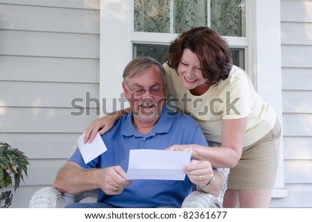 A middle aged couple reads a letter on the porch of their vintage house, enjoying reading good news together.