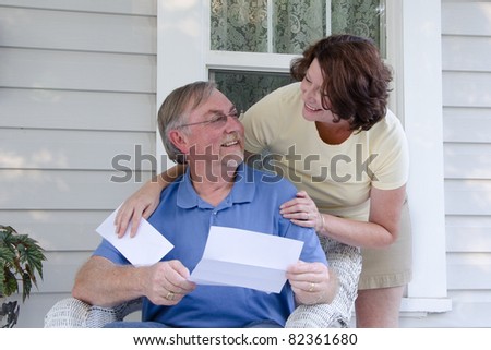 A middle-aged couple on an old-fashioned porch look at each other while reading a letter or announcement.