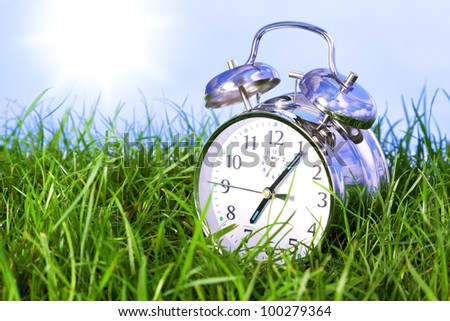 Photo of a chrome alarm clock outdoors sitting in grass on the morning of a bright sunny day.