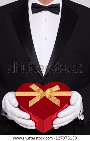 Man wearing black tie and white gloves holding a red heart shaped box of chocolates.