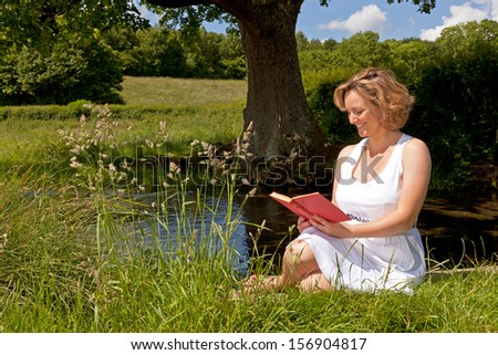 A woman in a white dress sat by a stream reading a book on a bright summers day.