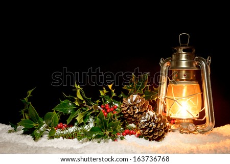 An oil filled lantern sitting in snow with holly, ivy and pine cones illuminated by the glow of the lamp.
