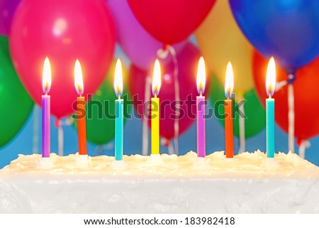 Candles burning on an white iced birthday cake with multicoloured balloons in the background, copy space on the cake to add your own message.