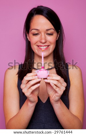 Happy brunette woman holding a small birthday cake with candle on pink background