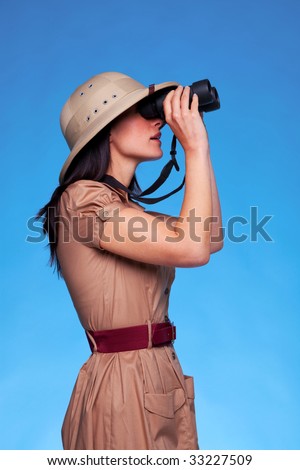 A woman wearing a pith helmet searching with a pair of binoculars, blue background with copy space.
