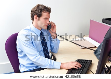 A male office worker answering the phone and looking at a computer monitor in a customer service center.