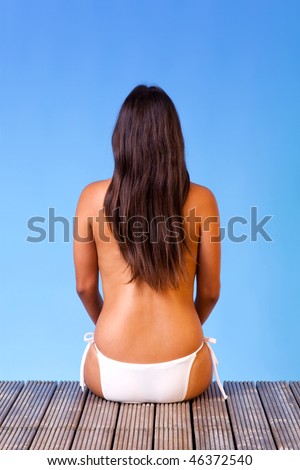 Rear view of a topless young woman wearing white bikini bottoms sat on the end of a wooden pier