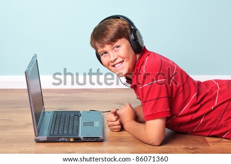 Photo of a boy wearing headphones looking towards camera lying on the floor next to his laptop computer.