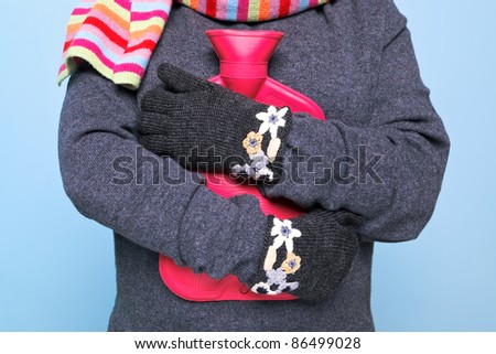 Photo of a woman holding a red hot water bottle to her chest whilst wearing hand kniitted woolen gloves trying to keep warm, good image for winter illness or warmth related themes.