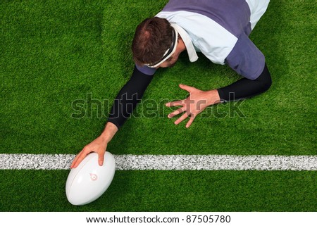 Overhead photo of a rugby player stretching over the line to score a try with one hand on the ball.