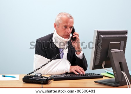 Photo of a mature businessman with injuries talking on the phone whilst trying to work on his computer. Good image for health and safety or accident at work related themes.
