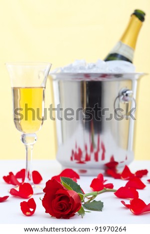 Photo of a red rose and a glass of champagne on a white linen tablecloth with a champagne bottle in an ice bucket in the background.
