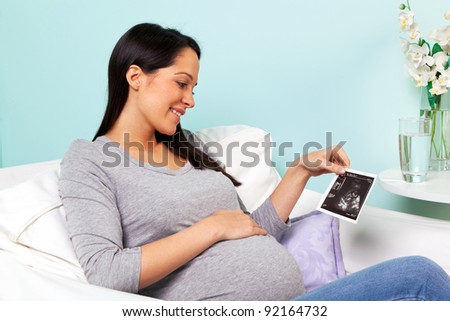 Photo of a pregnant woman at home sitting in an armchair looking at her baby ultrasound scan print.