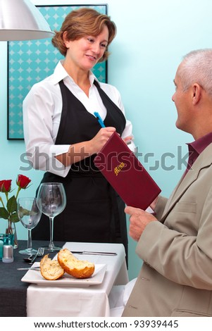Photo of a waitress in a restaurant taking a food order from a mature male who is sat at a table holding a menu.