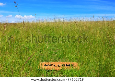 Photo of a welcome doormat in a grass meadow on a bright sunny day with blue sky and sunshine.
