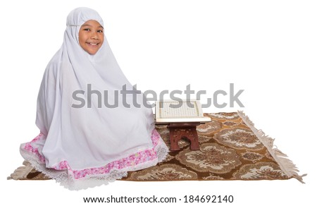 Young Asian Muslim girl in white hijab reading Al Quran on a prayer mat over white background.