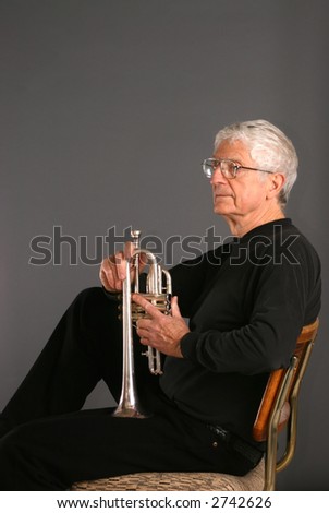 Old man sitting in a cane back chair holding a trumpet before a dark background