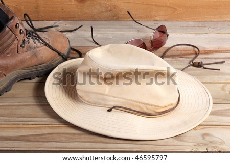Old fabric outdoor hat with leather strap and leather hiking boot with laces and dark glasses on a weathered wooden plank deck