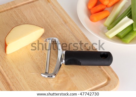 Gouda cheese with red rind on a bare wooden cutting board with a steel wire cutter cut celery and peeled carrots on a ceramic plate against a white background